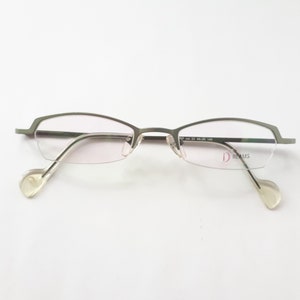 Könnte beinhalten: Silberfarbene, randlose Brille mit einer subtilen Biegung im Rahmen. Der Markenname "i DREAMS" ist in Pink auf der rechten Linse aufgedruckt.