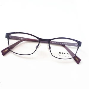 Peut inclure: Lunettes de vue violet foncé avec une monture rectangulaire et des branches bordeaux. Les lunettes présentent un motif subtil sur le haut de la monture et la marque "KLiiK denmark" est visible.