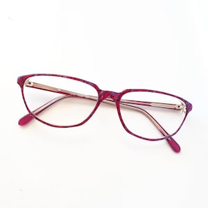 May include: A pair of eyeglasses with a burgundy and purple patterned frame. The lenses are clear, and the temples are a light gold color. The glasses are set against a white background.
