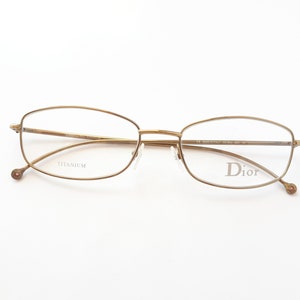 Könnte beinhalten: Goldfarbene Titan-Brille mit rechteckigem Gestell und dem Wort "Dior" auf dem rechten Bügel.
