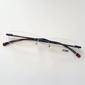 Könnte beinhalten: Eine Brille ohne Rahmen mit schwarzem Gestell und blauen Akzenten. Die Brille hat einen silbernen Nasensteg und der Markenname "STING EYEWEAR" ist auf der rechten Linse aufgedruckt.