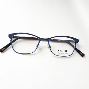 Könnte beinhalten: Blaue Brille mit Metallrahmen und rechteckiger Form. Die Brille hat einen dünnen Rahmen und der Markenname "KLiiK denmark" ist auf dem rechten Glas sichtbar. Der Rahmen misst 47/17-140.