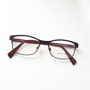 Peut inclure: Lunettes de vue en métal de couleur bordeaux avec une monture rectangulaire. Les verres sont transparents et le nom de la marque "KLIIK DENMARK" est imprimé sur la branche droite.