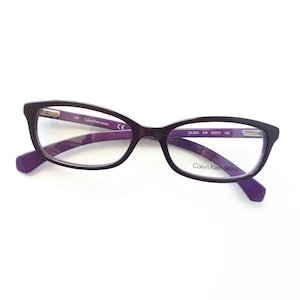 Könnte beinhalten: Lila und schwarze Brille mit einem Calvin Klein Jeans Logo am rechten Bügel. Die Brille hat eine rechteckige Form mit abgerundeten Ecken.
