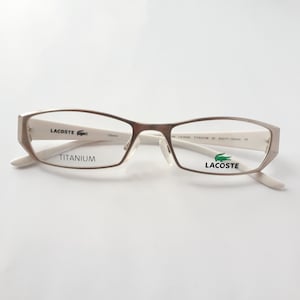 Könnte beinhalten: Weiße Titan-Gestellbrille mit einem grünen Krokodil-Logo am rechten Bügel und dem Wort "LACOSTE" auf dem rechten Bügel. Der linke Bügel trägt den Schriftzug "TITANIUM".