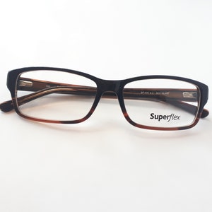 Könnte beinhalten: Braune und schwarze Schildpatt-Brille mit rechteckigem Gestell. Die Gläser sind klar und der Markenname "Superflex" ist auf dem rechten Glas aufgedruckt.