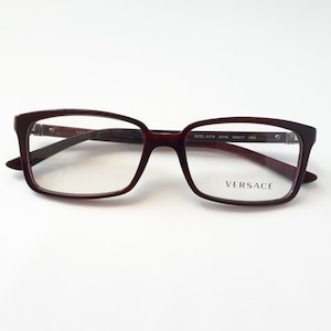 Könnte beinhalten: Burgunderfarbene rechteckige Brille mit einem schwarzen Versace-Logo auf der rechten Linse. Der Rahmen hat einen silbernen Akzent an den Bügeln. Der Text "MOD 3174 5045 55□17 145" ist auf dem rechten Bügel aufgedruckt.