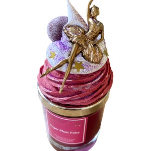 Puede incluir: Una vela aromática rosa y morada con una figurita de bailarina dorada en la parte superior. La vela está en un frasco de vidrio con una tapa plateada y la etiqueta dice "Sugar Plum Fairy".