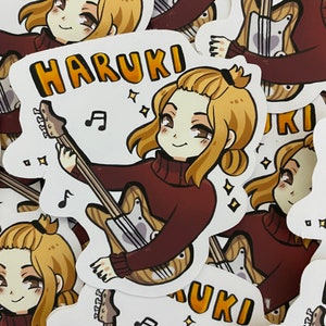 Puede incluir: Una pegatina con un personaje de dibujos animados con cabello rubio tocando una guitarra. El personaje lleva un suéter rojo y la pegatina tiene el nombre "HARUKI" escrito en letras amarillas.
