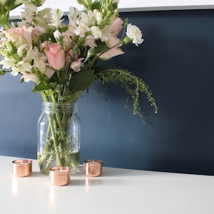 May include: A bouquet of pink and white flowers in a clear glass jar on a white surface with three copper tea light candle holders.