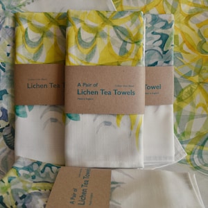 May include: A pair of white cotton tea towels featuring a yellow and green lichen design. The towels are packaged with a brown paper label that reads "A Pair of Lichen Tea Towels".