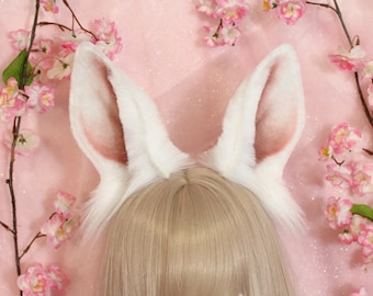 May include: A pair of tall, pointed animal ears made of white faux fur with soft pink shading on the inner ear surfaces. The ears are attached to a headband and are positioned above a light blonde wig, framed by branches with pink blossoms.