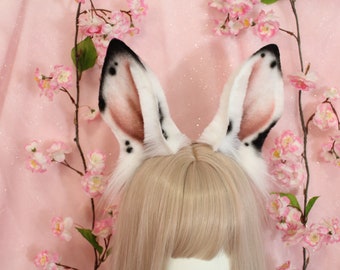 May include: A pair of tall, pointed faux fur animal ears featuring white fur with black tips and dark spots. The inner ear area is shaded with a soft pink hue, and the ears are attached to a headband worn over light blonde hair.