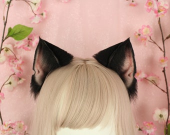 May include: A pair of black faux fur animal ears attached to a thin headband. The ears feature a pointed shape with black fur on the outer edges and a lighter, pinkish-toned interior texture, positioned atop a light blonde wig with straight hair.