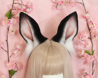 May include: A pair of tall, pointed animal ears made of faux fur, featuring a black outer edge that transitions into a white base. The inner ear is lined with a soft, light pink fabric. The ears are attached to a headband worn over blonde hair.