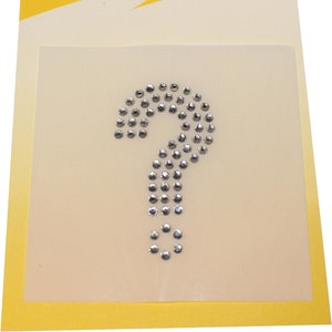 May include: A silver question mark made of rhinestones on a white background. The background is surrounded by a yellow border. The text "KLEIBER APPLIDUR" is printed in black on the yellow border.
