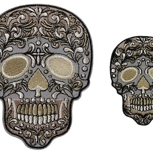 May include: Two embroidered sugar skull patches in gray, black, and gold. The larger skull is detailed with floral patterns and a spade design. The smaller skull is a simpler design.