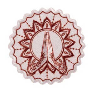 May include: A circular patch with a white background and a scalloped edge. The patch features a detailed brown design of hands in a prayer position, surrounded by a mandala-style floral pattern. The design is centered within a circle.