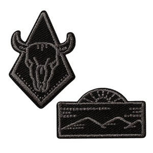 May include: Two black embroidered iron-on patches. The first patch is a diamond shape with a bull skull inside. The second patch is a rectangular shape with a mountain range and a sun.