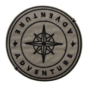 May include: Circular patch with the word "ADVENTURE" in black, arched around the outer edge of the patch. A black compass rose is in the center. The patch is a muted olive green color.
