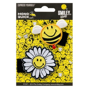May include: A pack of two iron-on patches featuring a smiling bee and a daisy with a smiley face. The patches are on a black background with a white border and a yellow smiley face pattern. The text "Express Yourself" and "Smiley World" are printed on the packaging.