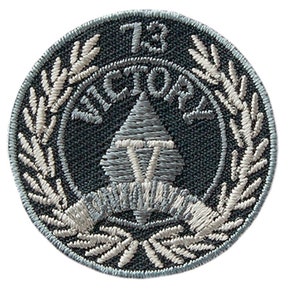 Puede incluir: Un parche bordado gris con una corona de laurel blanca, una punta de lanza gris con una "V" blanca en su interior y el texto "73rd VICTORY" en blanco.