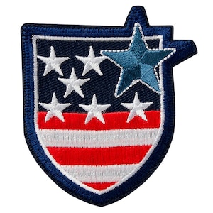 Könnte beinhalten: Gestickter Aufnäher mit einem Schilddesign und blauem Rand. Der Aufnäher zeigt weiße Sterne auf marineblauem Hintergrund, rot-weiße Streifen und einen blauen Stern oben. Er ist für Kleidung oder Accessoires bestimmt.