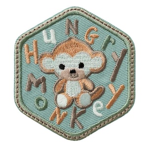Könnte beinhalten: Gestickter Aufnäher in sechseckiger Form mit helltürkisfarbenem Hintergrund und braunem Rand. Der Aufnäher zeigt einen Cartoon-Affen mit den Worten "HUNGRY MONKEY" in verschiedenen Farben.