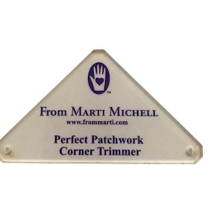 Può includere: Uno strumento triangolare in plastica trasparente con un logo a mano viola e il testo "From Marti Michell" e "www.frommarti.com". Il testo "Perfect Patchwork Corner Trimmer" è anche stampato sullo strumento.