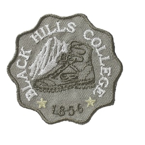 Könnte beinhalten: Ein grauer gestickter Aufnäher mit dem Text "Black Hills College" und einem Stiefel mit einem Berg im Hintergrund. Das Jahr "1886" ist ebenfalls im Design enthalten.