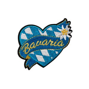 Könnte beinhalten: Ein blauer und weißer herzförmiger Patch mit einer weißen Edelweißblume und einem blauen Band mit dem Wort "Bayern" in Gold gestickt.