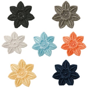 Op de afbeelding: Een verzameling van zeven geborduurde bloemvormige patches in verschillende kleuren. De patches zijn zwart, grijs, wit, lichtblauw, oranje, geel en marineblauw. Elke patch heeft een gedetailleerd bloemdesign met acht bloemblaadjes.