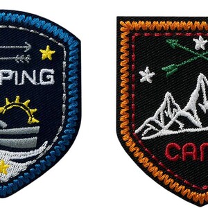 Könnte beinhalten: Zwei gestickte Patches mit Camping-Motiv. Der erste Patch ist blau mit weißen Nähten und zeigt eine Person in einem Boot mit dem Wort "CAMPING" darüber. Der zweite Patch ist schwarz mit orangenen Nähten und zeigt eine Bergkette mit dem Wort "CAMP" darunter.