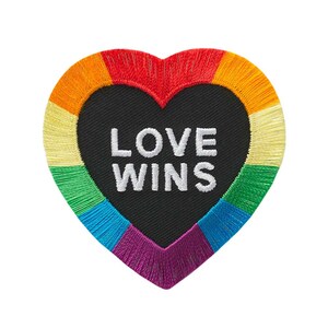 Pode incluir: Um remendo em forma de coração arco-íris com o texto "LOVE WINS" em bordado branco sobre fundo preto.