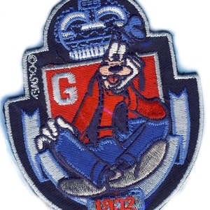 May include: An embroidered patch featuring a cartoon dog wearing a blue hat and sitting on a red shield with a white letter G. The patch has a blue crown at the top and the year 1972 at the bottom.