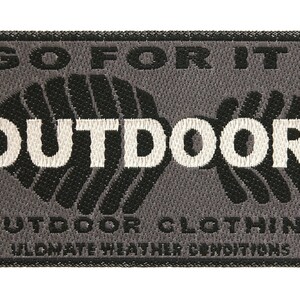 以下が含まれることがあります： 黒と白の刺繍が施されたグレーの織りパッチ。パッチには「Go For It! Outdoor Outdoor Clothing Ultimate Weather Conditions」と書いてあります。