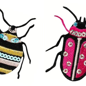 May include: Two embroidered insect patches. One is black with yellow stripes and teal sequins. The other is pink with a white center and silver sequins. Both have black antennae and legs.