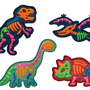 Peut inclure: Quatre patchs thermocollants colorés représentant des squelettes de dinosaures. Les patchs sont de différentes couleurs, notamment bleu, vert, rouge et orange. Les dinosaures sont un T-Rex, un dinosaure au long cou, un dinosaure volant et un dinosaure à trois cornes.