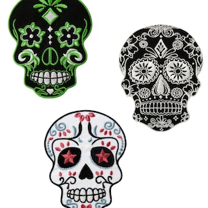 Op de afbeelding: Drie geborduurde suiker schedel patches. Eén is zwart met witte en groene details, één is wit met zwarte en roze details en één is zwart met witte details.