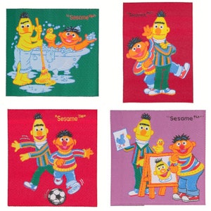 Puede incluir: Cuatro parches de tela coloridos con personajes de Barrio Sésamo. Uno muestra a Beto limpiando una bañera con Enrique y un patito de goma. Otros muestran a Beto y Enrique jugando, con el logo de Barrio Sésamo.