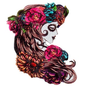 Puede incluir: Un parche bordado colorido que presenta a una mujer con una corona de flores y cabello largo y fluido. El rostro de la mujer está pintado con un diseño de calavera, un motivo común en la cultura mexicana. El parche es perfecto para agregar un toque de fantasía y tradición a la ropa, las bolsas u otros accesorios.