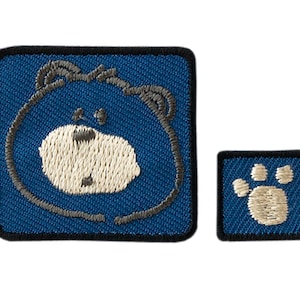 Op de afbeelding: Twee geborduurde strijkpatches. Eén met een blauw beren gezicht met een witte neus en een voetafdruk. De andere patch is een blauwe voetafdruk met een wit midden.