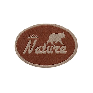 Könnte beinhalten: Ein brauner, ovaler gestickter Aufnäher mit dem Wort "Nature" in beigefarbener Schrift und einer beigefarbenen Silhouette eines Bären, der vor einer Bergkette geht.