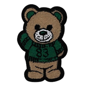 Könnte beinhalten: Ein brauner Teddybär, der einen grünen Pullover mit der Nummer 83 trägt. Der Bär hat einen schwarzen Umriss und weiße Augen.