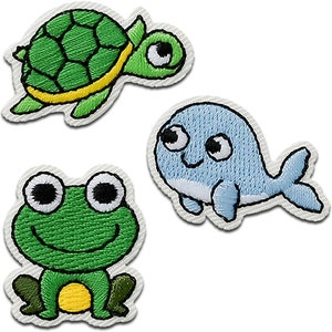 Könnte beinhalten: Drei gestickte Bügelbilder mit einem grünen Frosch, einer grünen Schildkröte und einem blauen Wal. Jedes Bügelbild hat große, weiße Augen und einen lächelnden Mund.