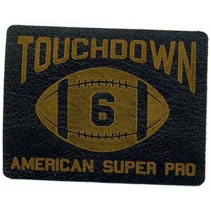 Könnte beinhalten: Ein schwarzer Leder-Patch mit goldener Beschriftung, die "TOUCHDOWN" und "AMERICAN SUPER PRO" lautet. In der Mitte des Patches befindet sich ein goldener Football mit der Nummer "6".