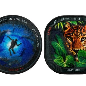 May include: Two round camera lens-shaped patches. The left patch has a blue background with a silhouette of a scuba diver surrounded by fish. The text "DEEP IN THE SEA ZOOM LENS 1:3.5-5.6" is printed around the edge. The right patch has a green background with a close-up of a leopard's face. The text "LENS EF 35mm 1:1.8 CAPTURE" is printed around the edge.