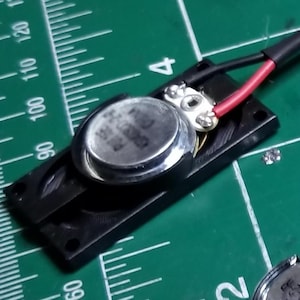 May include: A small, silver-colored speaker component mounted on a black plastic base. Red and black wires are connected to the speaker. The background is a green cutting mat with measurement markings.