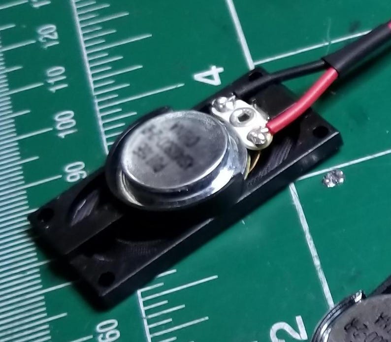 May include: A small, silver-colored speaker component mounted on a black plastic base. Red and black wires are connected to the speaker. The background is a green cutting mat with measurement markings.