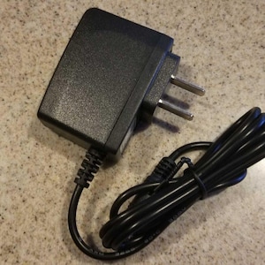 May include: A black AC power adapter with a two-prong plug and a black cord. The adapter is rectangular with a textured surface. The cord is coiled and secured.
